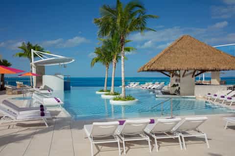 Infinity pool overlooking ocean with palm trees, white lounge chairs, purple accents, thatched-roof cabana, and clear blue sky