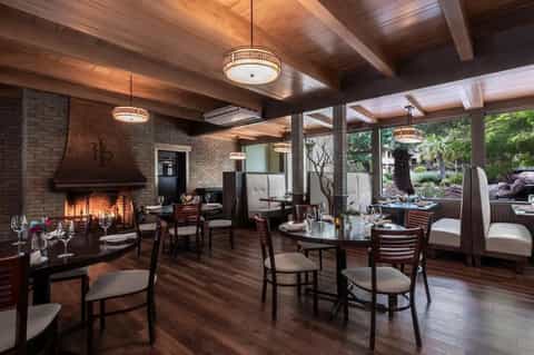 Upscale restaurant interior with exposed brick fireplace, wooden beams, dark furniture, and glass doors