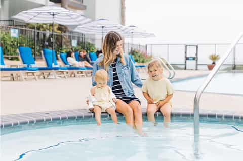 Woman and two toddlers at edge of shallow pool with umbrellas and lounge chairs visible in sunny resort setting
