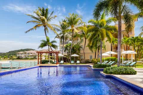 Beachfront resort pool with blue water, palm trees, and yellow building facade