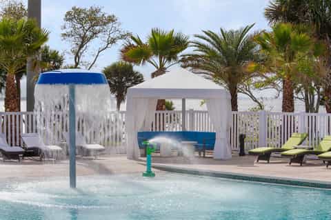Resort pool area with bucket shower, white cabanas, palm trees, and lounge chairs overlooking waterfront