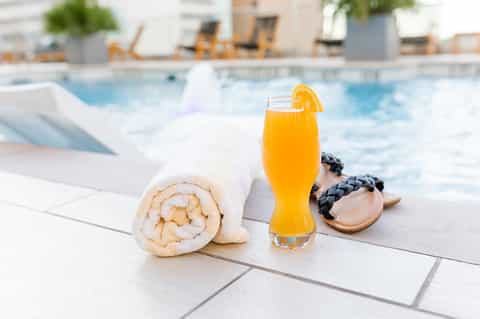 Poolside refreshment with rolled white towel, orange juice, and accessories on white pavers