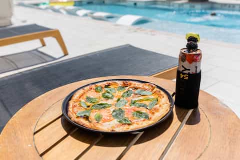 Gourmet pizza with basil leaves on wooden table beside resort pool with lounge chairs