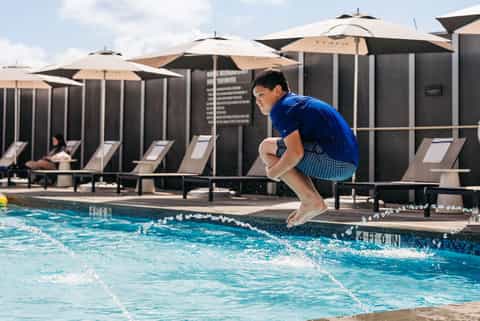 Man jumping into pool at resort with black cabanas, tan umbrellas, and loungers in background