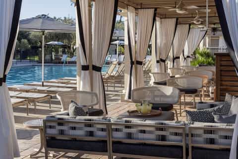 Covered poolside cabanas with white curtains, lounge chairs, and view of resort pool