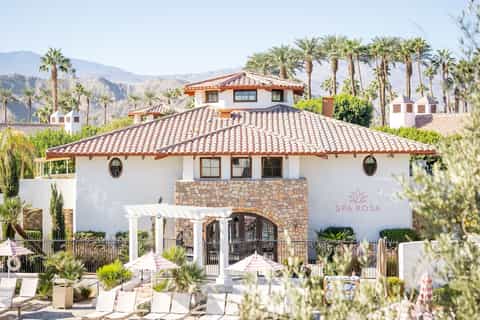 Spa Rosa building with Spanish tile roof, stone entrance, white pergola, palm trees, and mountain backdrop