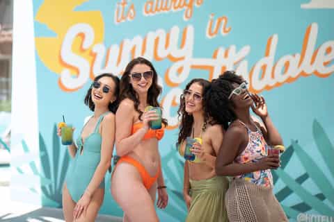 Group of women in swimwear posing by turquoise wall with 'Sunny Hayday' text at pool area