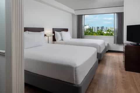Modern hotel room with two queen beds, windows overlooking city skyline and residential buildings