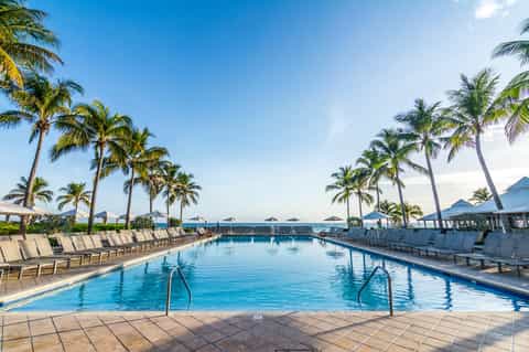 Resort pool with palm trees, lounge chairs, umbrellas, and ocean view in background