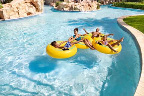 Resort lazy river with families floating on yellow inflatables, rock formations, and lush green landscaping
