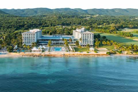 Beachfront resort aerial view showing white buildings, pools, golf course, mountains and turquoise ocean