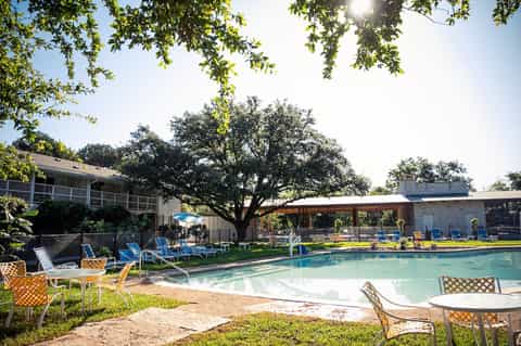 Resort pool area with loungers, shade structures, mature oak trees, and stone buildings on sunny day