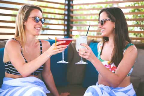 Two women in bikinis toasting colorful cocktails under wooden pergola