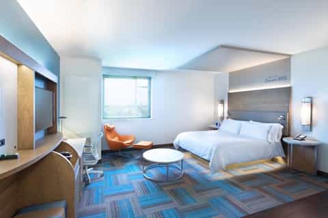 Spacious hotel suite with bed, lounge area, desk, orange chair, and floor-to-ceiling windows