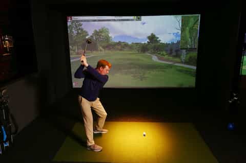 Man using golf simulator with large screen display showing virtual golf course gameplay