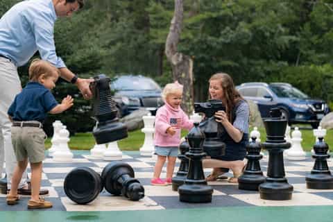 Family playing giant outdoor chess game in resort courtyard with parking lot visible in background
