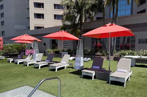 Resort lawn with white lounge chairs under red umbrellas, modern building, palm trees, and green grass
