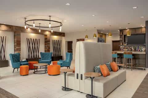 Modern hotel lobby lounge with turquoise chairs, orange ottomans, bar area, and pendant lighting