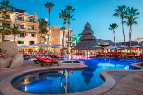 Resort pool at dusk with illuminated water, lounge chairs, tiki bar, palm trees, and hotel buildings