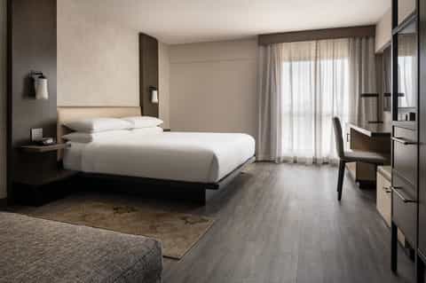 Modern hotel bedroom with white bedding, gray wood flooring, desk, and balcony with sheer curtains