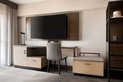 Hotel room desk with TV, work chair, shelving, and window with neutral decor