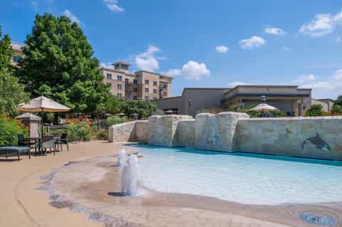 Resort pool with wave features and stone waterfall backdrop, surrounded by umbrellas and hotel buildings