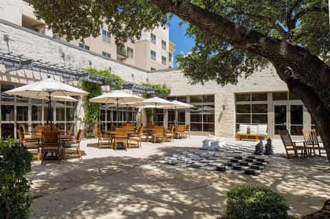 Stone patio courtyard with wooden seating, white umbrellas, and large tree providing shade