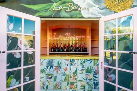 La Boisson bar with tropical botanical mural, pink doors, patterned tile counter, and bottle display