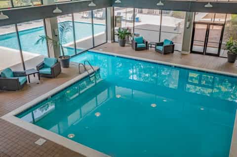 Indoor heated pool with turquoise water, lounge seating, and floor-to-ceiling windows