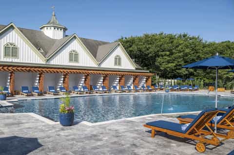 Resort pool with covered pavilion, blue umbrellas, wooden lounge chairs, and white building