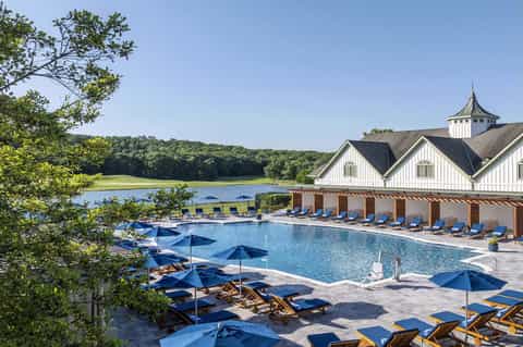 Resort pool with blue umbrellas, wooden loungers, and lakeside pavilion with white architecture