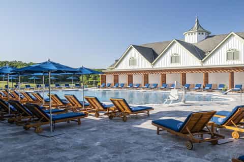 Resort pool with blue lounge chairs, umbrellas, and colonial-style pool house