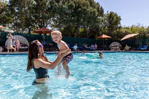 Family enjoying community pool with umbrellas, lounge chairs, and tree-lined surroundings