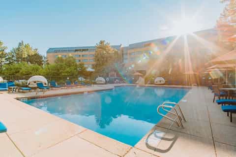 Resort pool at sunset with blue umbrellas, white lounge chairs, hotel building, and dramatic golden sunlight