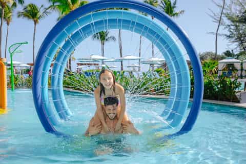 Father and daughter playing in pool through large blue tunnel toy with palm trees and umbrellas