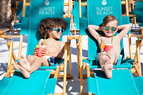 Two children relaxing in turquoise Sunset Beach lounge chairs with tropical drinks