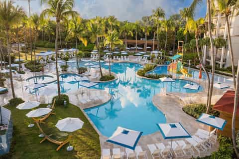 Large resort pool complex with palm trees, white lounge chairs, umbrellas, landscaping, and tropical setting
