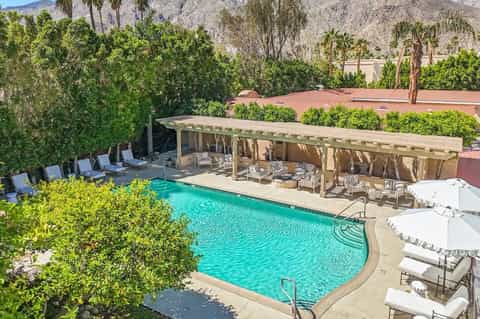Desert resort pool with wooden pergola shade structure, lounge chairs, mountains in background, and lush landscaping