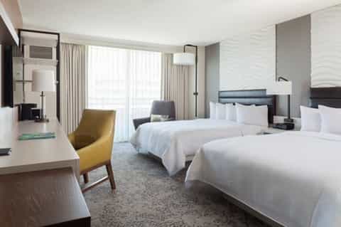 Modern hotel room with two beds, seating area, large windows, and neutral tones