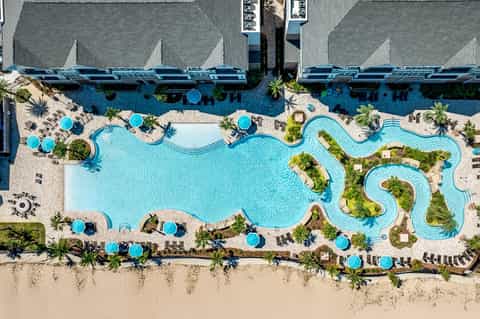 Aerial view of curved resort pool with blue umbrellas and lounge chairs along the beach