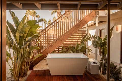 Outdoor bathroom with freestanding white bathtub, wooden stairs, and tropical plants on beach