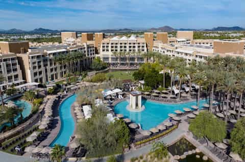 Aerial resort view showing curved blue pools, tan buildings, palm trees, and desert mountain landscape