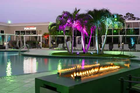 Nighttime pool area with colorful uplighting on palm trees, fire pit, and modern resort building