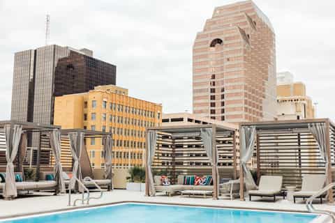 Urban rooftop pool area with cabanas, gray curtains, colorful pillows, and city skyline with tall buildings