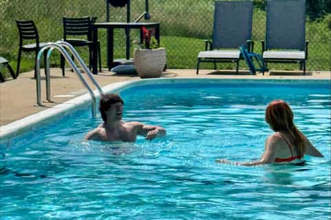 Guests swimming in outdoor pool with ladder, lounge chairs, and privacy fence