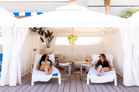 Two women lounging in white cabana beds under flowing white curtains and palm umbrella