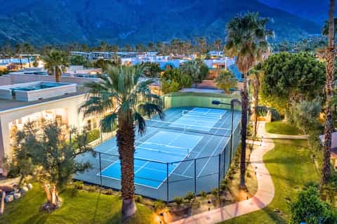 Illuminated tennis courts at dusk with palm trees, resort buildings, and mountain backdrop in background.