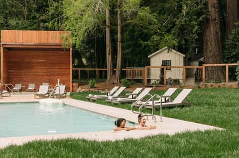 Guests relaxing in forest-surrounded pool with modern wood cabana and lounge chairs on grass