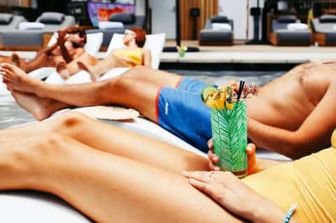 Guest holding tropical cocktail by pool while relaxing on lounge chairs at resort.