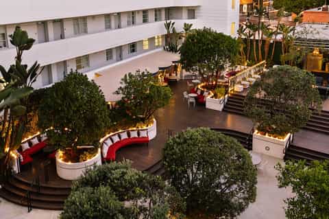 Modern hotel courtyard at dusk with curved red seating areas, palm trees, planters, and lit white building facade